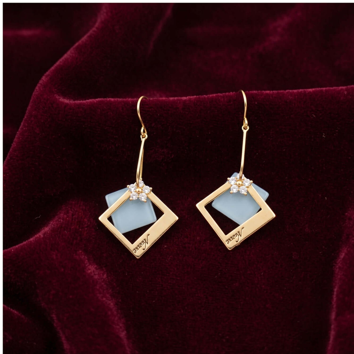 Golden square Floral Hoop earrings featuring blue stone and floral design on a plush burgundy background.
